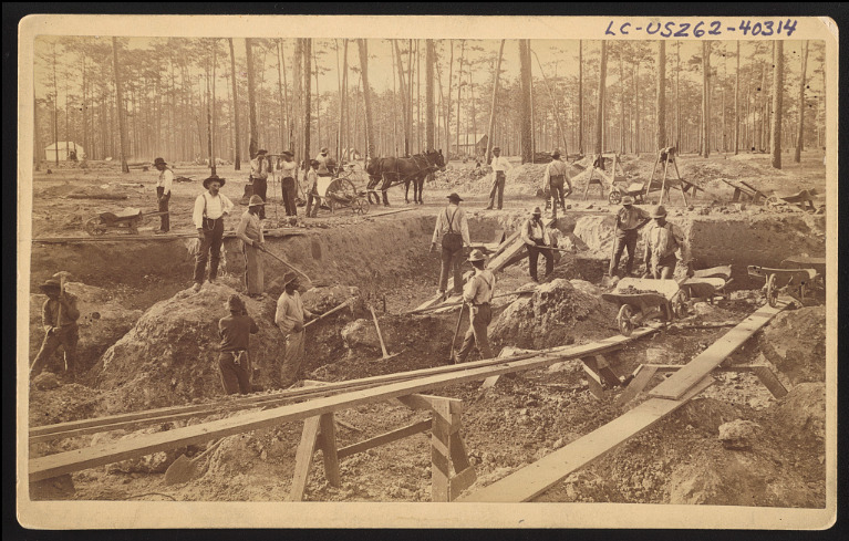 African American phosphate miners push wheelbarrows across thin wooden beams and swing pickaxes in a shallow pit phosphate mine in Dunnellon, Florida.